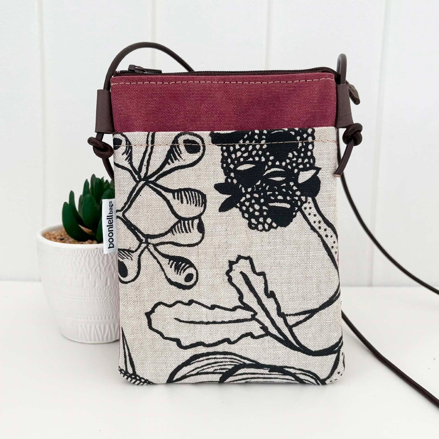 A small crossbody bag with a black and white botanical print on a linen-textured base, featuring a dusty rose top panel and dark brown straps. A small white tag on the side reads 'bookishbag'.