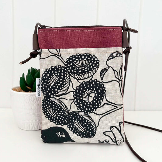 A small crossbody bag with a black and white botanical pattern on a linen-textured fabric, featuring a solid burgundy top panel and a dark brown leather strap, displayed next to a small potted succulent.